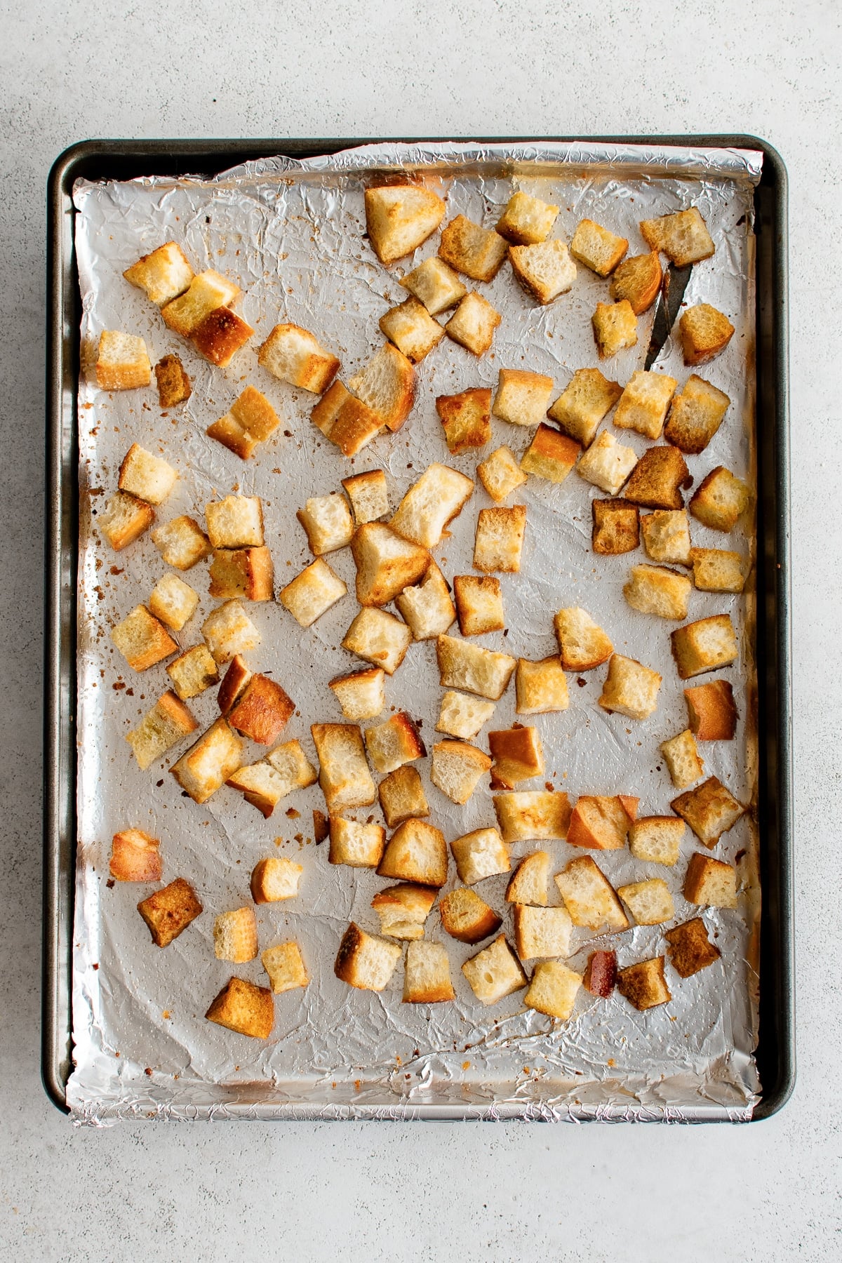 Overhead view of golden-brown homemade croutons spread across a foil-lined black rimmed baking sheet, with crisp toasted edges and scattered crumbs.