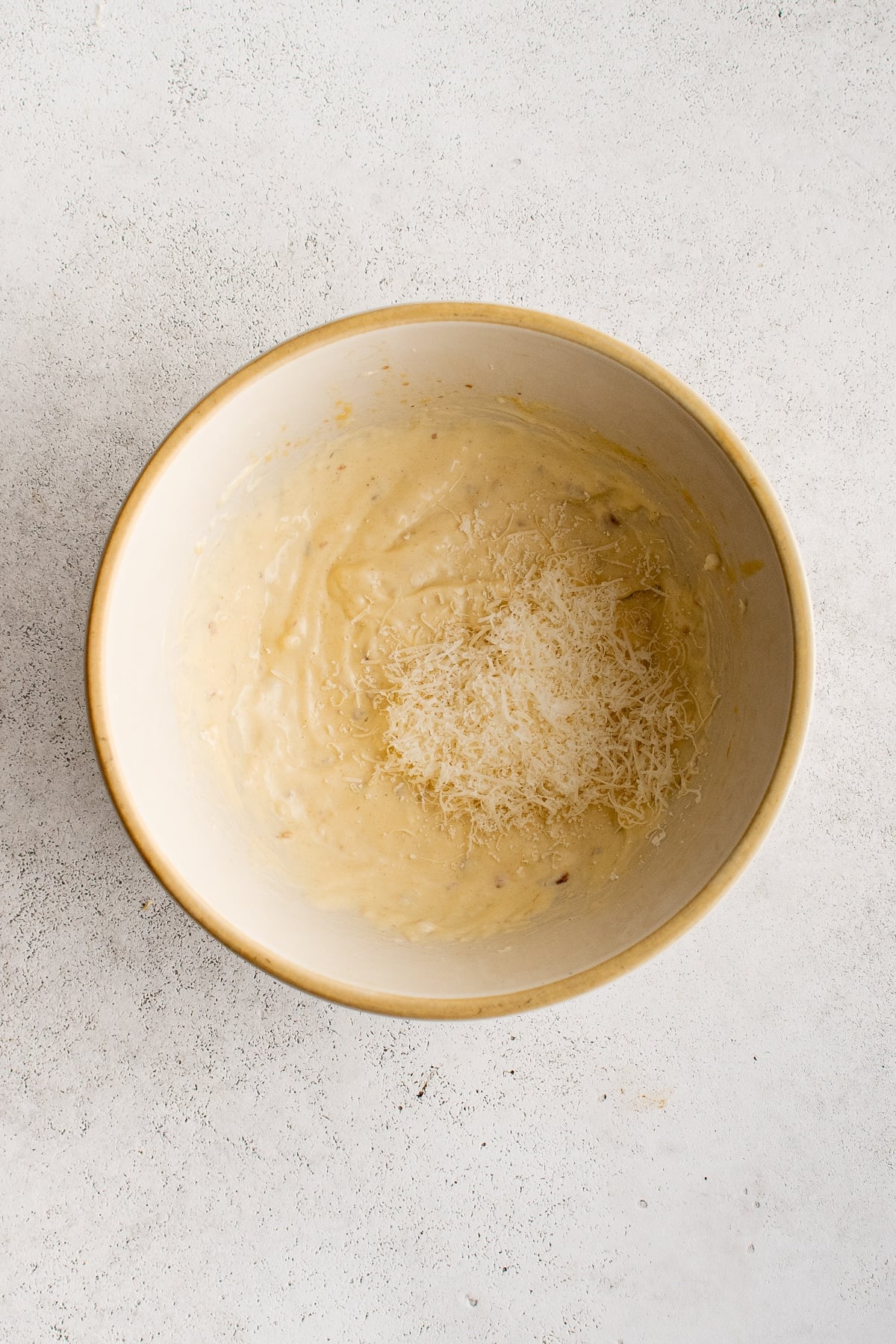 Overhead view of creamy homemade Caesar dressing in a cream-colored ceramic bowl with a tan rim, topped with a mound of finely grated Parmesan cheese on a light gray textured surface.