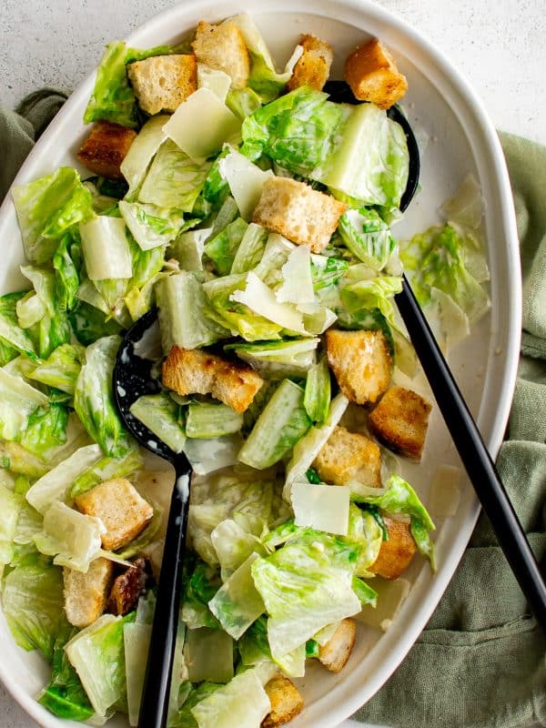 Overhead view of homemade Caesar salad in a large white oval serving platter with crisp green romaine, creamy dressing, golden croutons, and shaved Parmesan cheese. Two black serving utensils rest inside the salad, and a sage green cloth napkin is arranged around the right side of the platter.