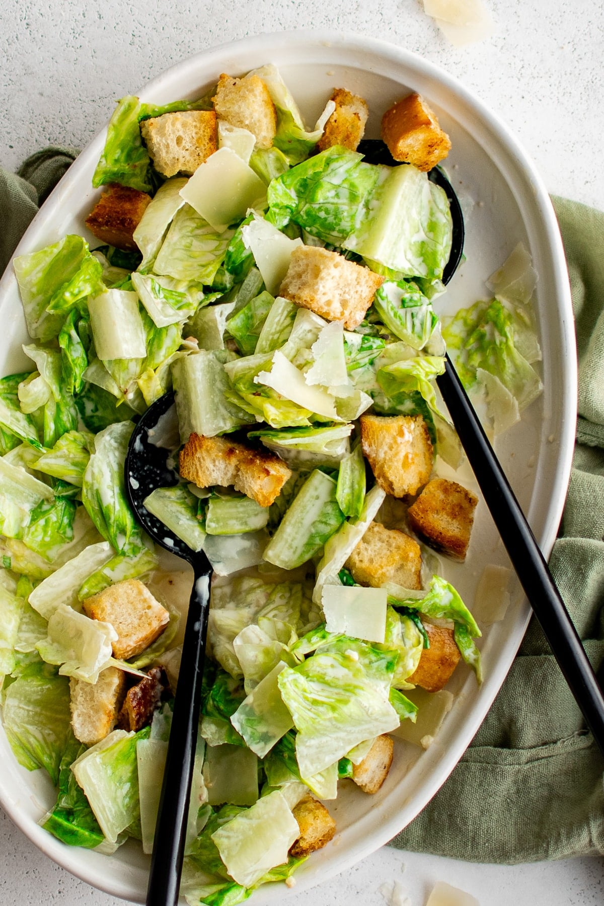 Overhead view of homemade Caesar salad in a large white oval serving platter with crisp green romaine, creamy dressing, golden croutons, and shaved Parmesan cheese. Two black serving utensils rest inside the salad, and a sage green cloth napkin is arranged around the right side of the platter.