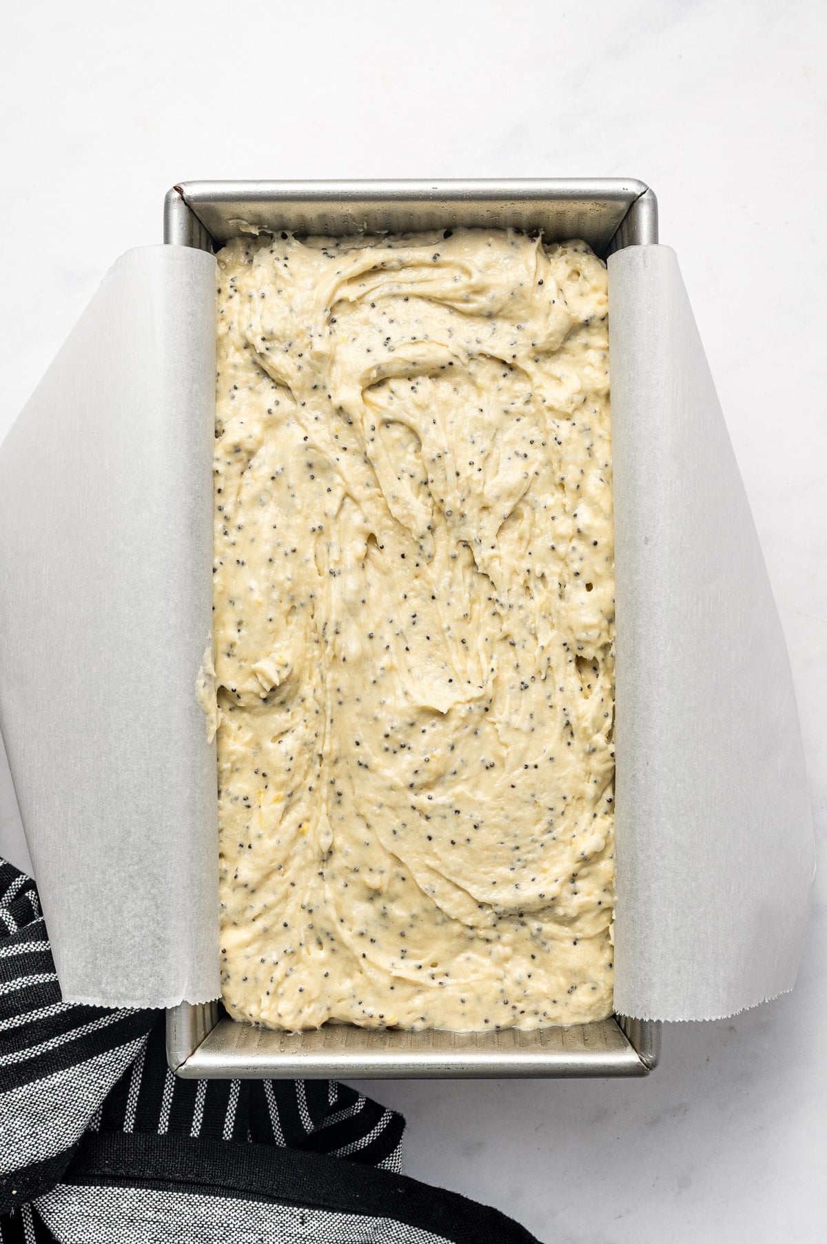 Overhead view of pale yellow lemon poppy seed batter spread inside a rectangular silver metal loaf pan lined with white parchment paper. Swirls from the spatula are visible across the surface, with a black-and-white striped towel at the lower left.