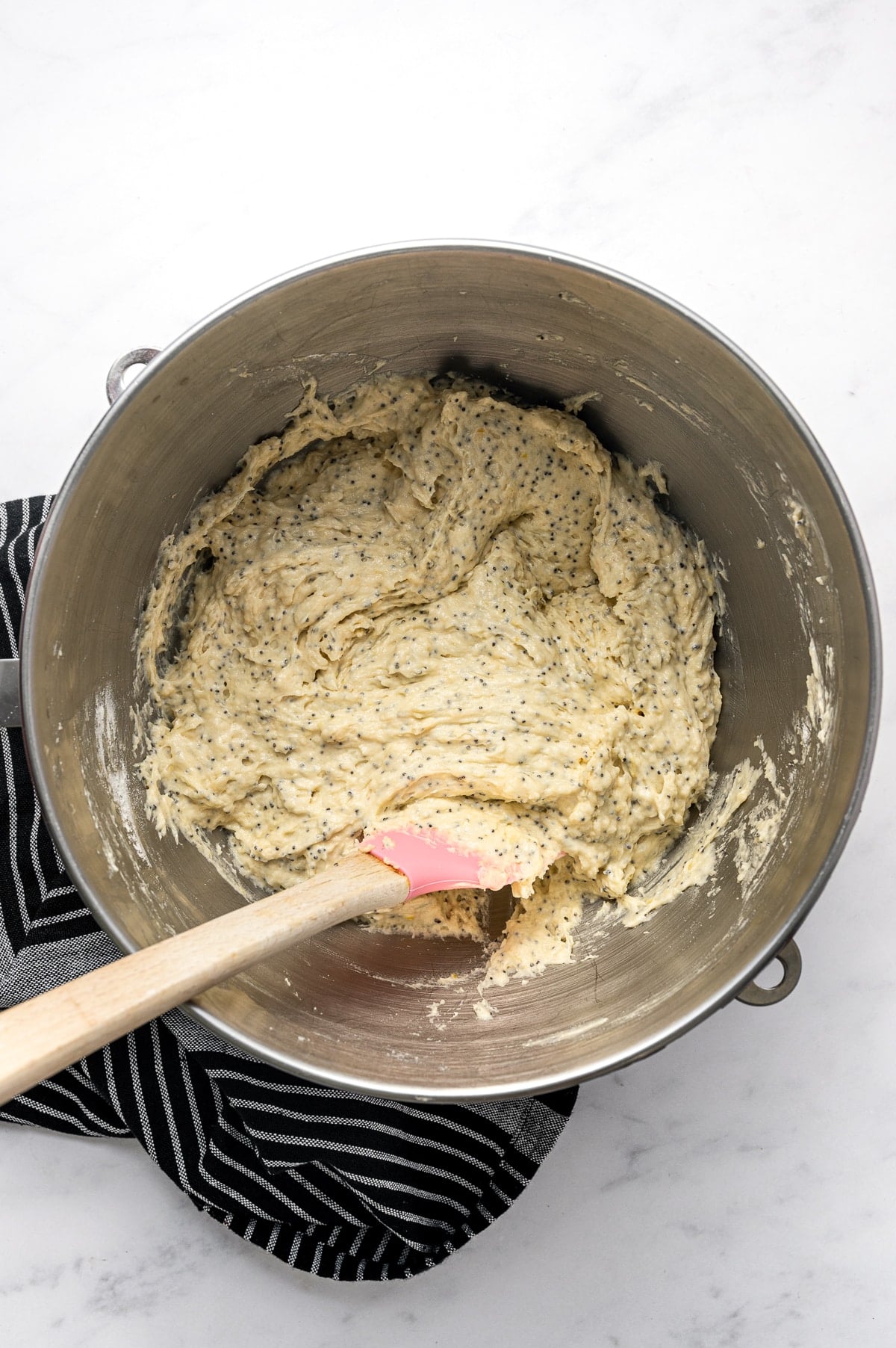 Overhead view of thick, pale yellow lemon bread batter evenly speckled with black poppy seeds inside a large stainless steel mixer bowl. A pink silicone spatula with a wooden handle rests in the batter, and a black-and-white striped towel is tucked beneath the bowl.