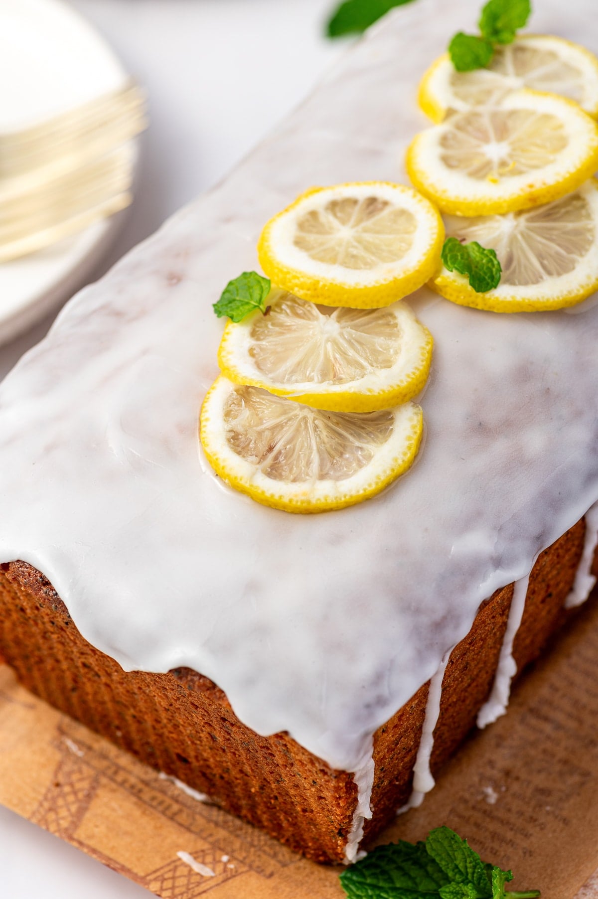 Close-up view of a golden-brown lemon poppy seed loaf covered with smooth white lemon glaze that drips down the sides. Several thin yellow lemon slices and small green mint leaves are arranged across the top, with a white plate and gold fork softly blurred in the background.