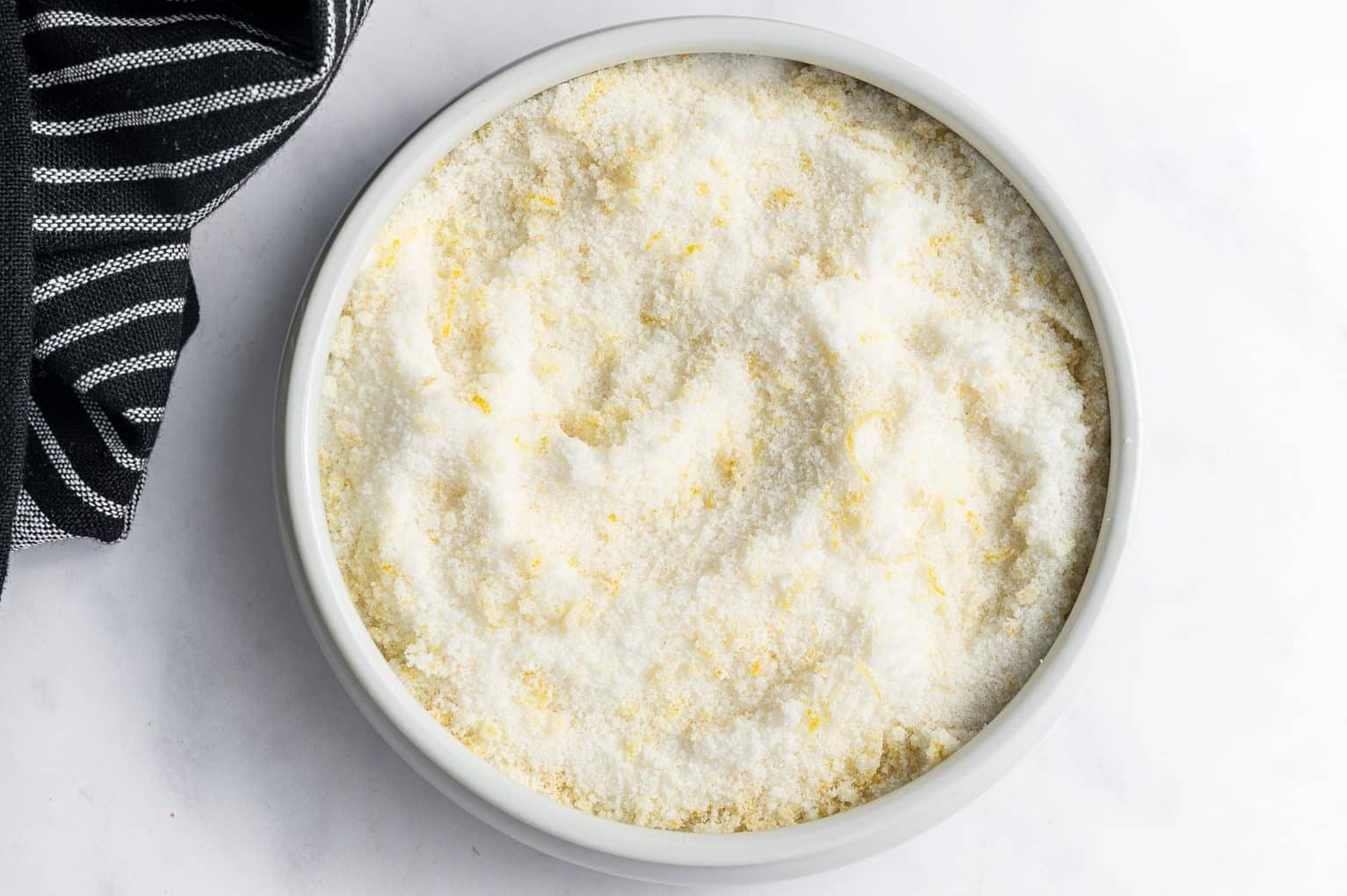 Overhead view of granulated sugar mixed with fine yellow lemon zest in a round white ceramic bowl on a white marble surface, with a folded black-and-white striped kitchen towel in the lower-left corner.