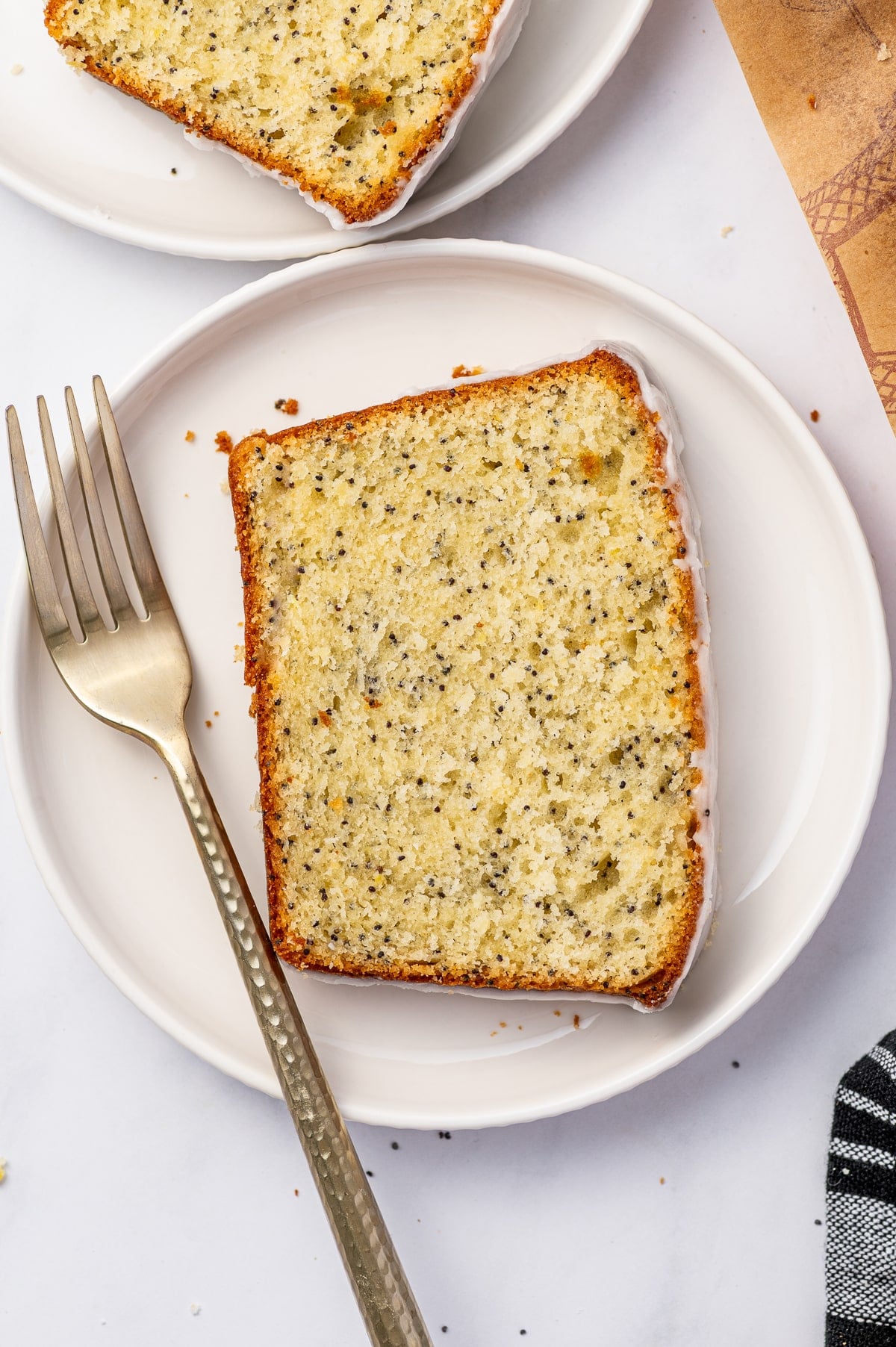 Overhead view of a thick slice of pale yellow lemon poppy seed bread on a round white scalloped plate, showing a moist crumb evenly speckled with black poppy seeds and a thin layer of white glaze. A gold fork rests to the left, with a second slice, scattered crumbs, a brown serving board, and a black-and-white striped towel around the plate.