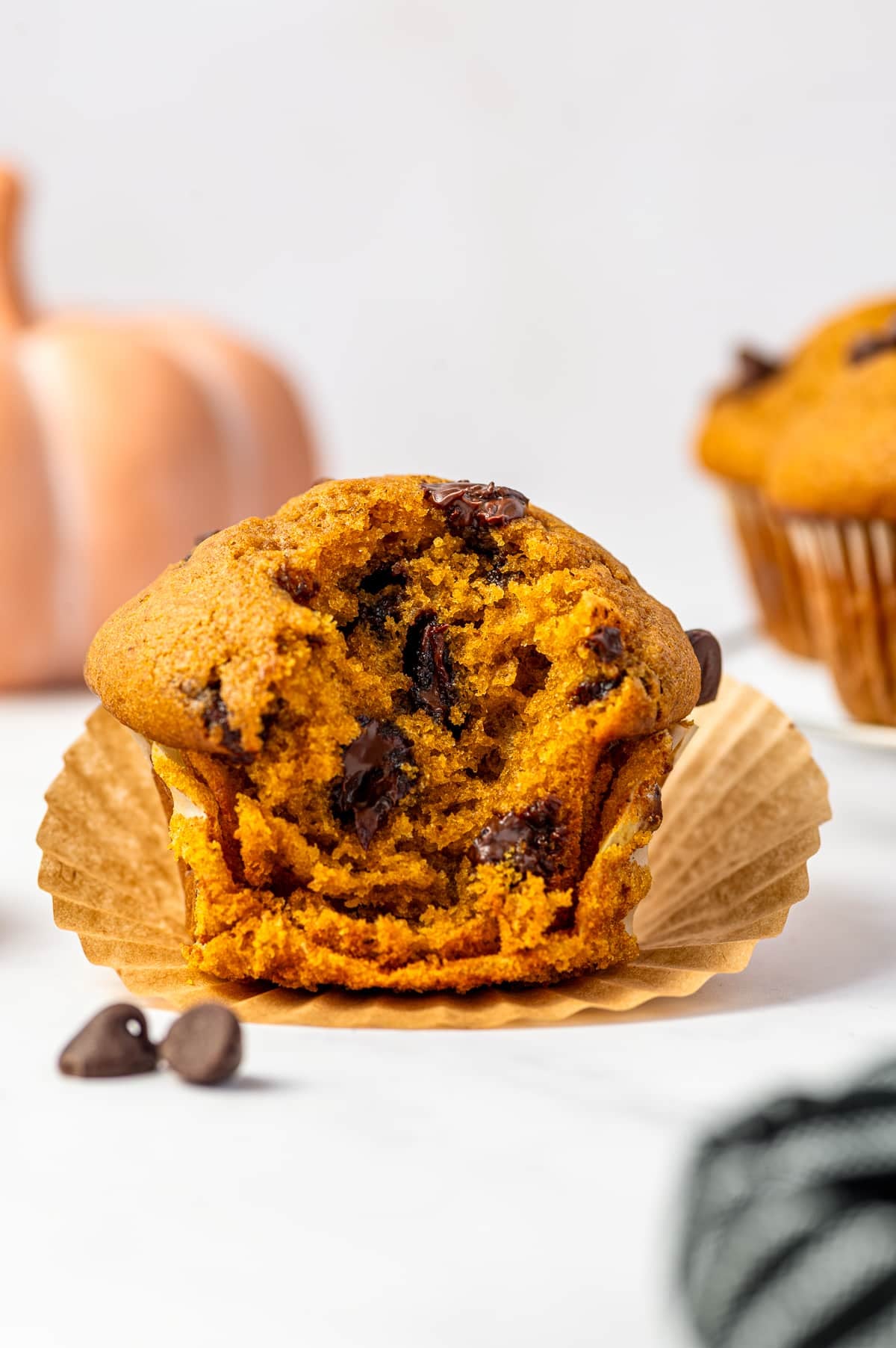 A pumpkin chocolate chip muffin broken open in its brown paper liner to reveal a soft orange crumb and pockets of melted chocolate, with two loose chocolate chips in front, a pale pink pumpkin on the left, another muffin blurred in the background, and a black-and-white striped towel at the lower right.