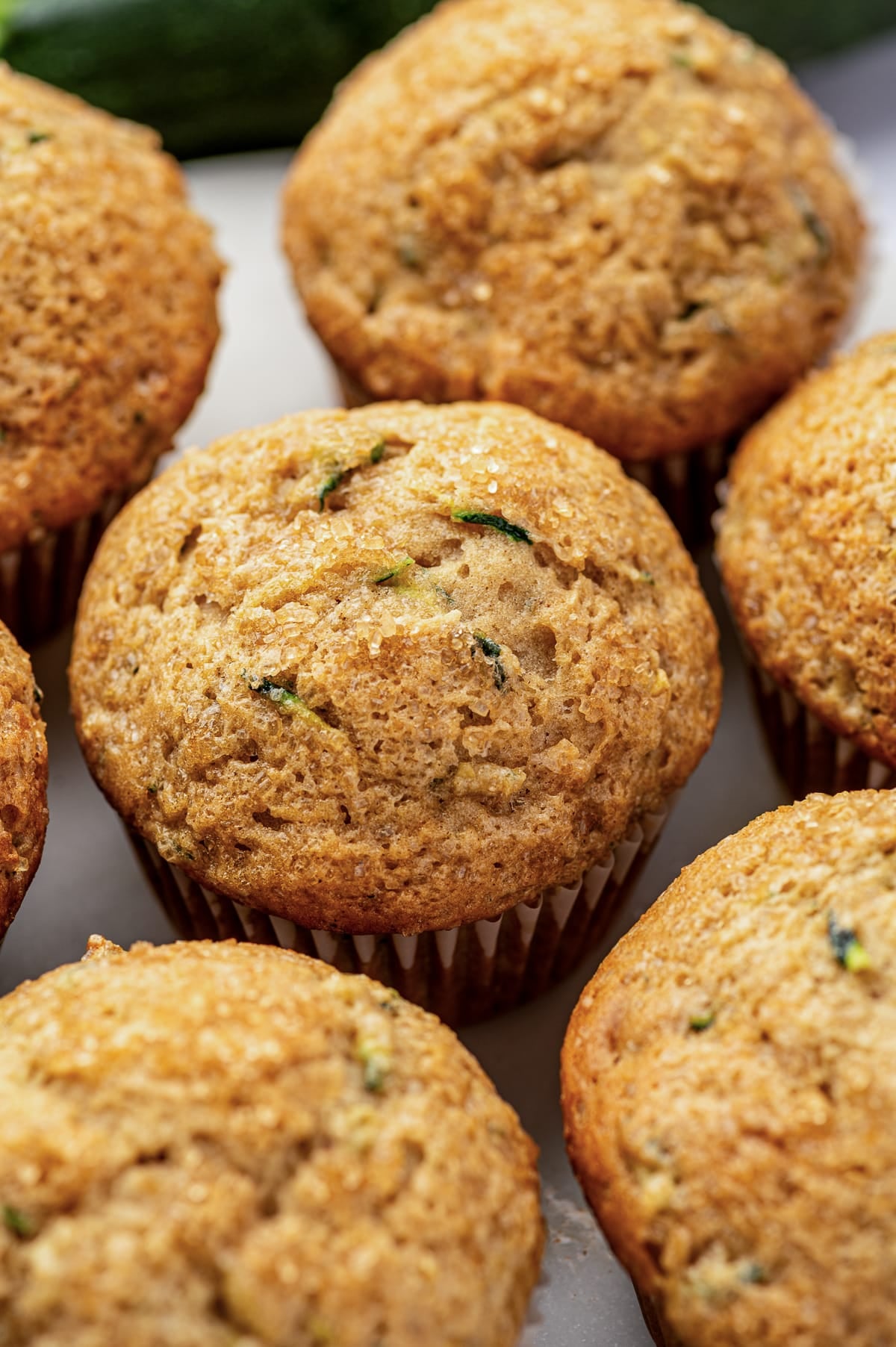 Close-up of a golden zucchini muffin with a tall rounded top, visible green zucchini flecks, and sparkling sugar crystals, surrounded by several additional muffins with whole dark green zucchini softly blurred in the background.