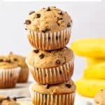 Vertical Pinterest graphic showing three golden banana protein muffins with mini chocolate chips stacked on a white plate, with additional muffins and yellow bananas in the background. Text reads "Protein Banana Muffins," "with vanilla protein powder," and "THEFORKEDSPOON.COM."
