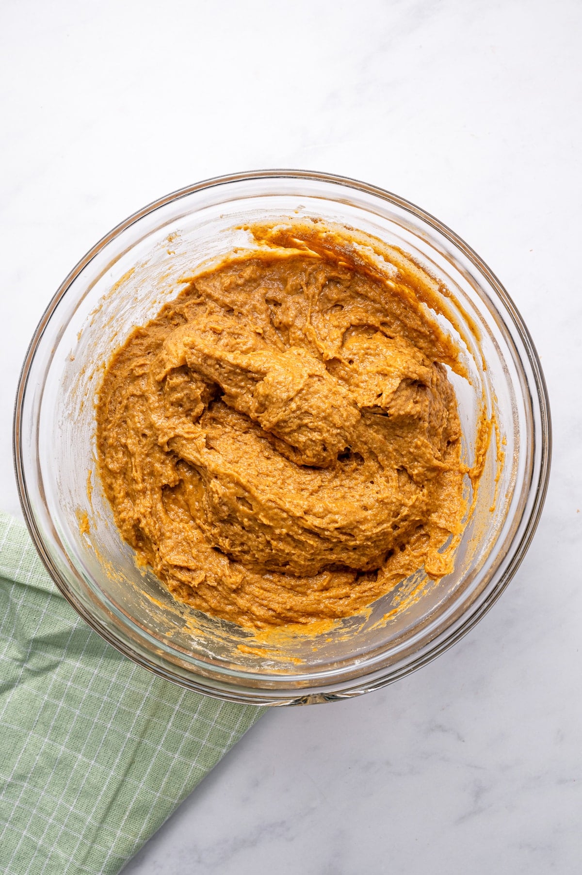 Thick orange-brown protein pumpkin muffin batter mixed in a large clear glass bowl, with visible swirls from folding and a green striped kitchen towel beside the bowl.