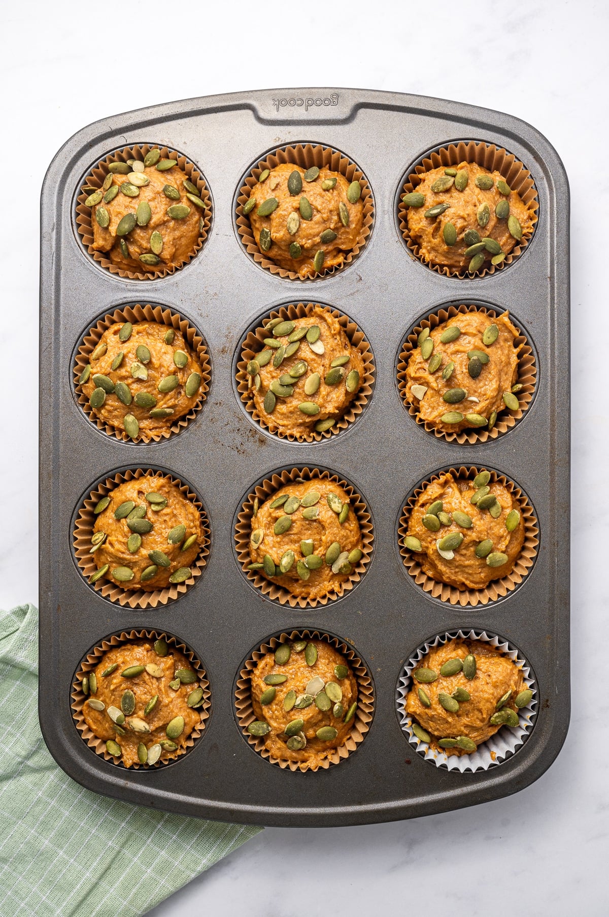 Twelve unbaked protein pumpkin muffins in a dark gray metal muffin pan, filled with thick orange pumpkin batter and generously topped with green pepitas. Most muffins are in brown paper liners, with one white liner in the bottom right cup, and a green striped towel is visible beside the pan.
