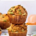 Pinterest graphic featuring two stacked golden-brown protein pumpkin muffins in brown paper liners topped with green pepitas. A white bowl of orange pumpkin puree, a small white bowl of pepitas, and a pale orange pumpkin appear in the background. Black text at the top reads "Protein Pumpkin Muffins" with smaller text underneath reading "with vanilla protein powder," and "THEFORKEDSPOON.COM" appears at the bottom.