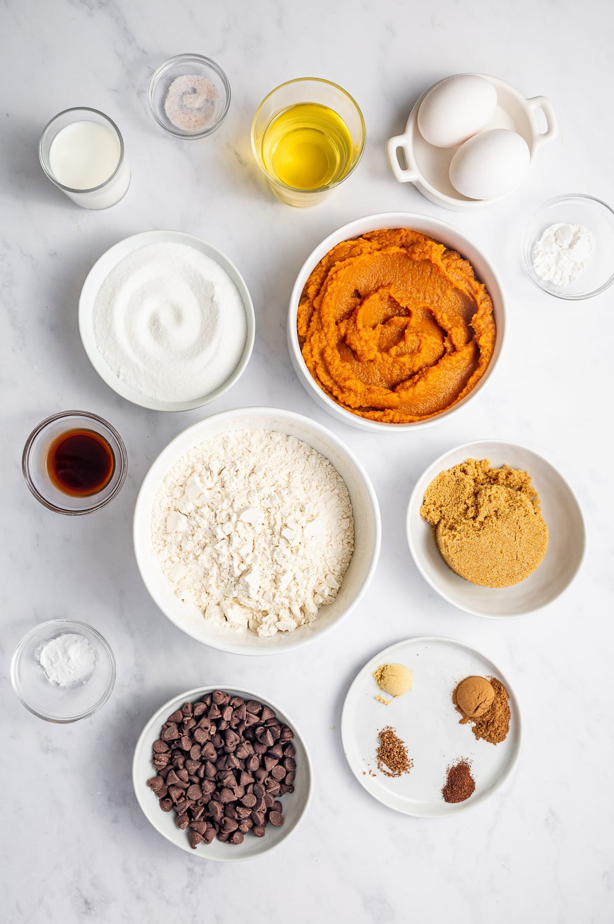 Overhead arrangement of ingredients for pumpkin chocolate chip muffins on a white marble surface, including white bowls of all-purpose flour, granulated sugar, light brown sugar, pumpkin puree, semi-sweet chocolate chips, warm spices, baking powder and baking soda, plus two white eggs, neutral oil, whole milk, vanilla extract, and salt.