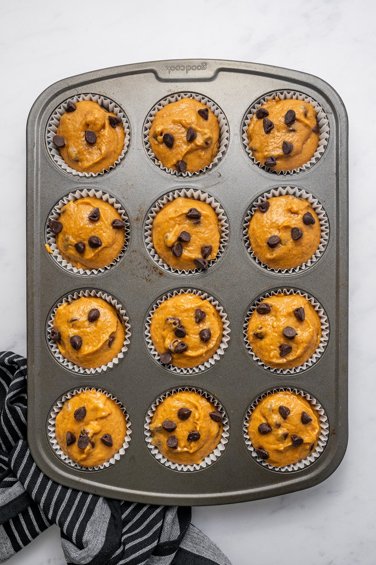 Twelve paper-lined muffin cups filled nearly to the top with orange pumpkin muffin batter and sprinkled with semi-sweet chocolate chips in a gray metal muffin pan, photographed overhead on a white marble surface with a black-and-white striped towel.