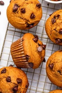 Several golden-orange pumpkin chocolate chip muffins cooling on a metal wire rack over a white surface, including one muffin resting on its side in a brown paper liner, with semi-sweet chocolate chips scattered across the muffin tops and surface.