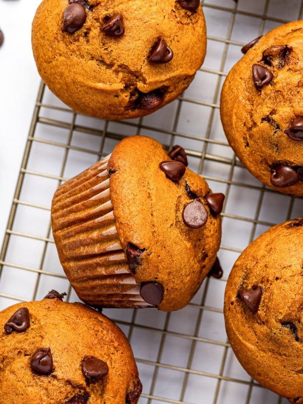 Several golden-orange pumpkin chocolate chip muffins cooling on a metal wire rack over a white surface, including one muffin resting on its side in a brown paper liner, with semi-sweet chocolate chips scattered across the muffin tops and surface.