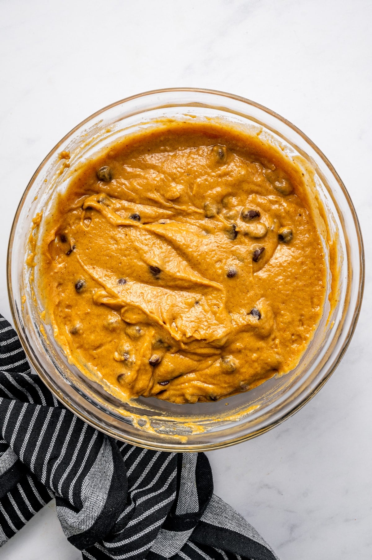 Thick orange pumpkin muffin batter with semi-sweet chocolate chips folded throughout in a large clear glass mixing bowl, photographed overhead on a white marble surface with a black-and-white striped kitchen towel along the lower left side.
