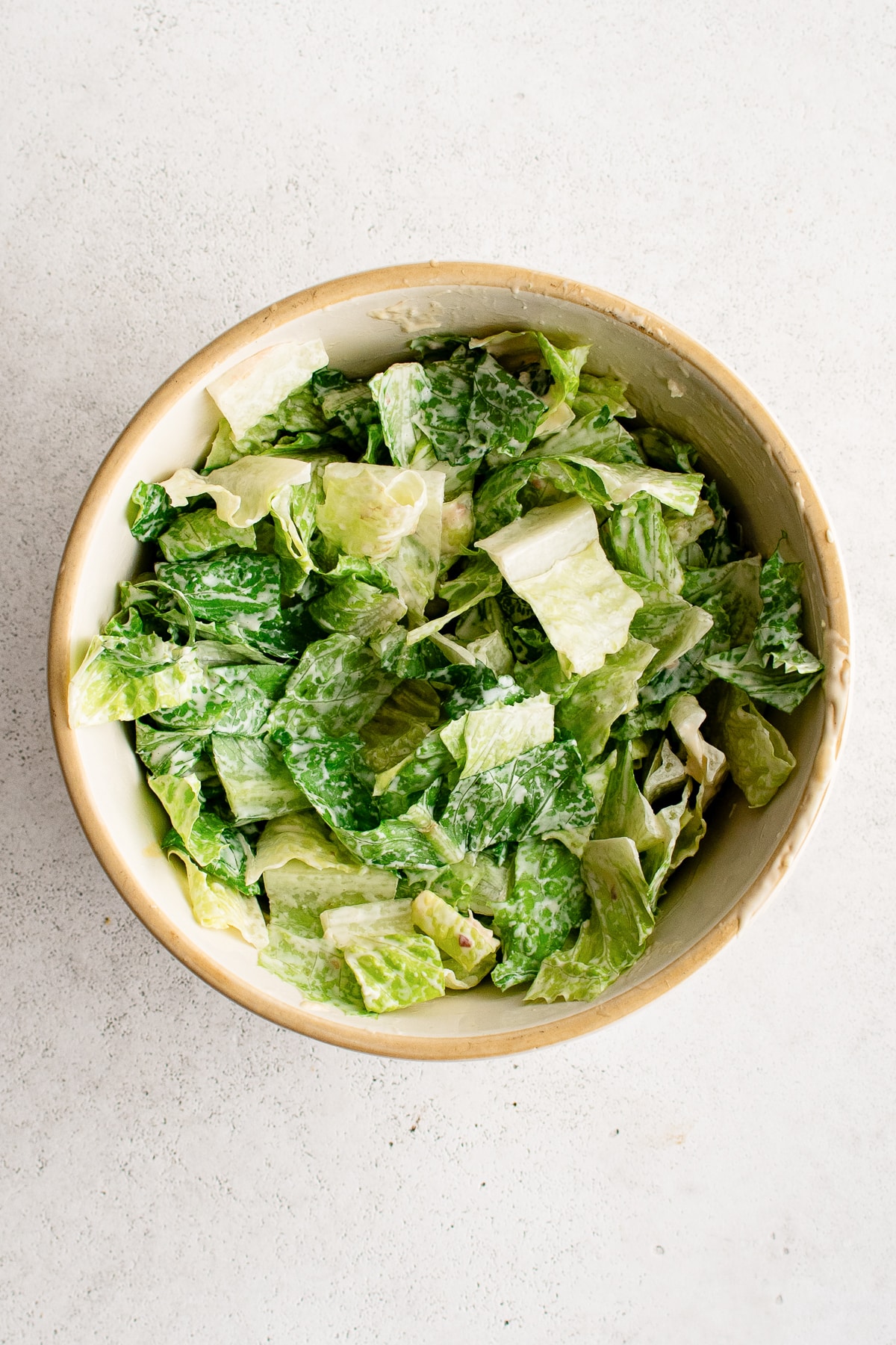 Overhead view of chopped green romaine lettuce coated in creamy Caesar dressing inside a cream-colored ceramic mixing bowl with a tan rim on a light gray textured surface.