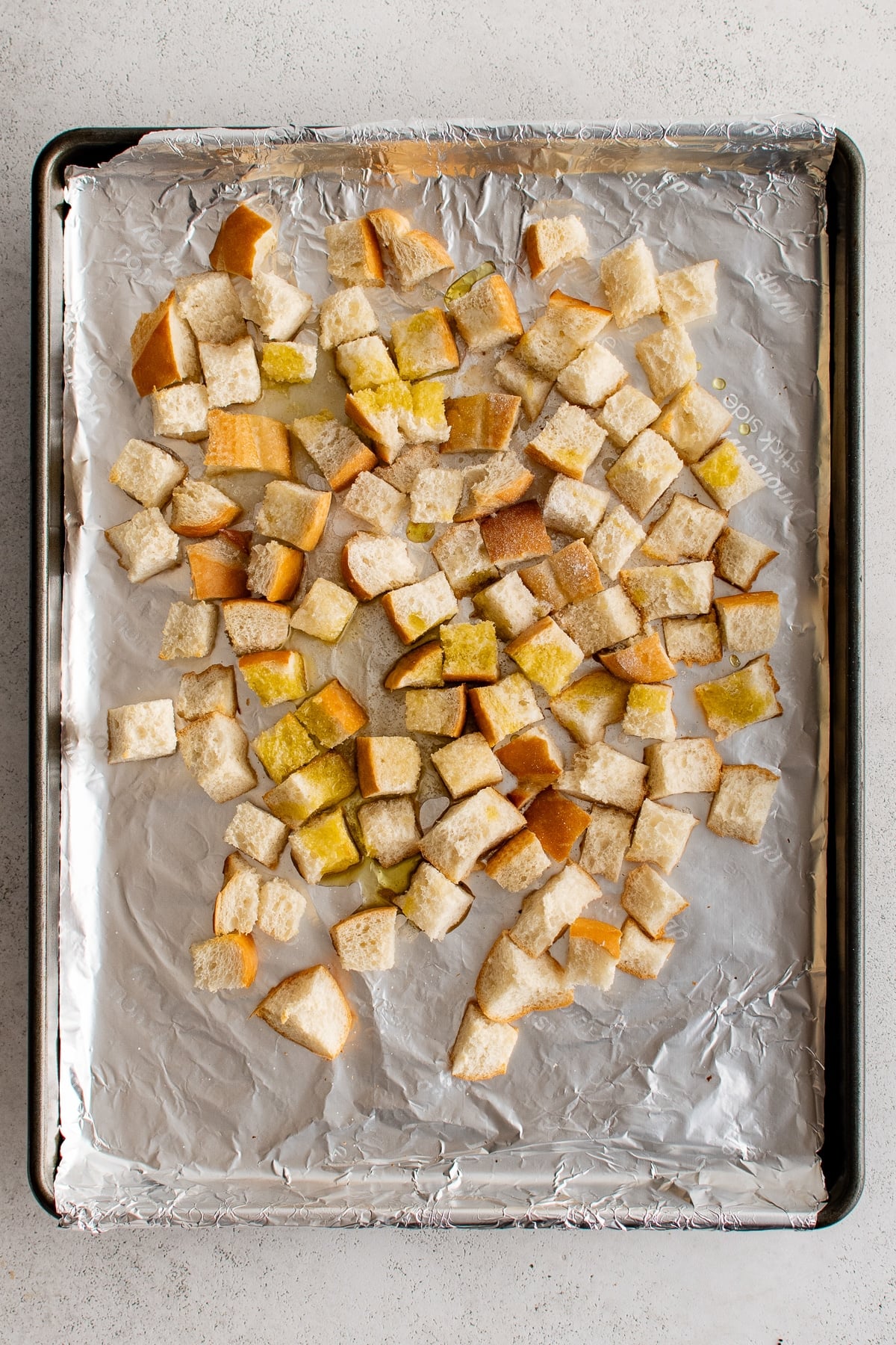 Overhead view of cubed French bread spread across a foil-lined black rimmed baking sheet and drizzled with golden olive oil before baking into homemade croutons.