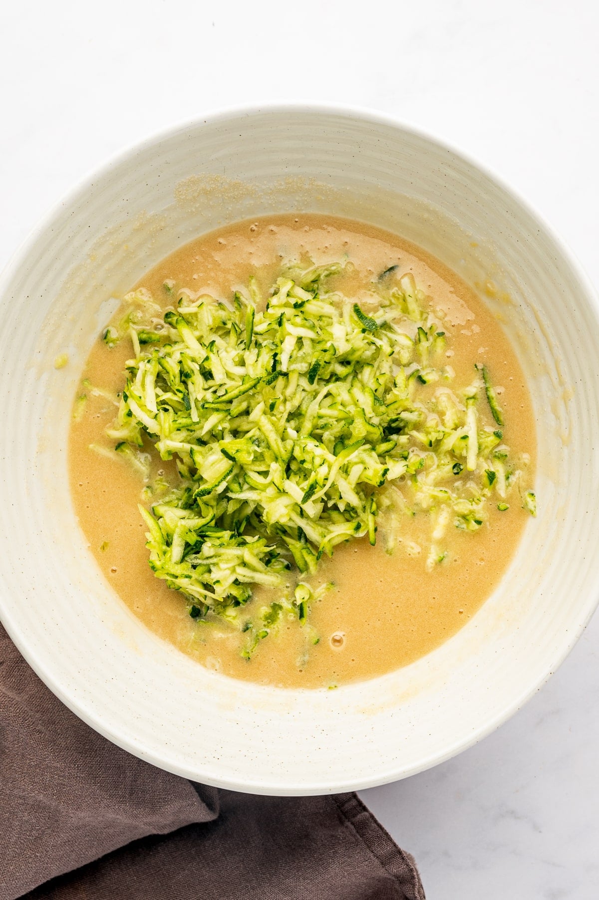 Overhead view of finely shredded bright green zucchini piled in the center of a tan wet muffin batter in a large off-white ceramic mixing bowl. A brown cloth napkin is partially visible beside the bowl on a light countertop.