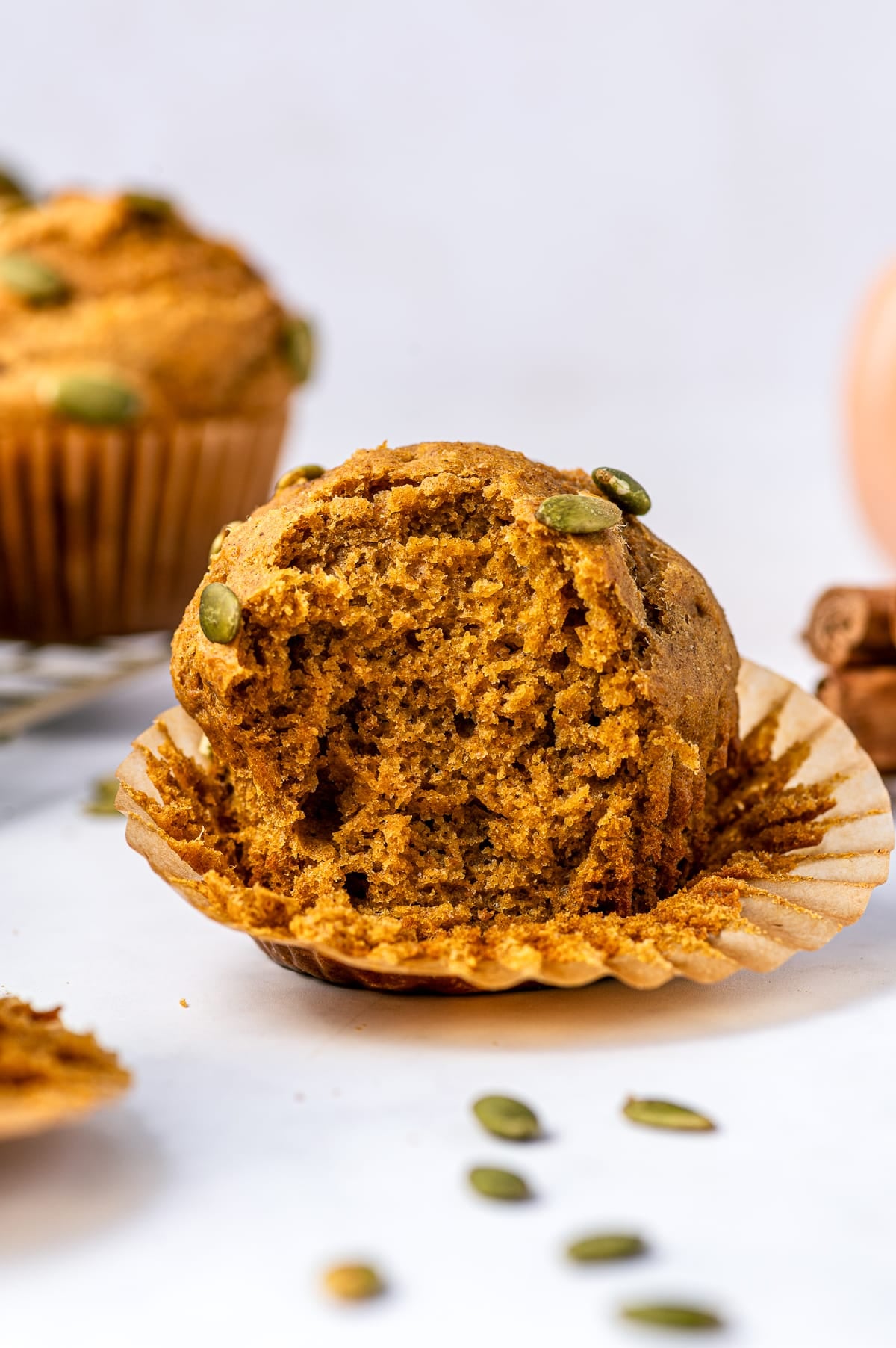 A protein pumpkin muffin broken open to reveal a soft, fluffy orange-brown crumb, with its brown paper liner peeled back. Green pepitas sit on top of the muffin, with another muffin and fall baking ingredients softly blurred in the background.
