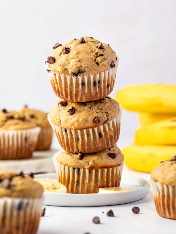 Three golden banana protein muffins with mini chocolate chips stacked on a white plate, surrounded by additional muffins, banana slices, scattered chocolate chips, and whole yellow bananas in the background.