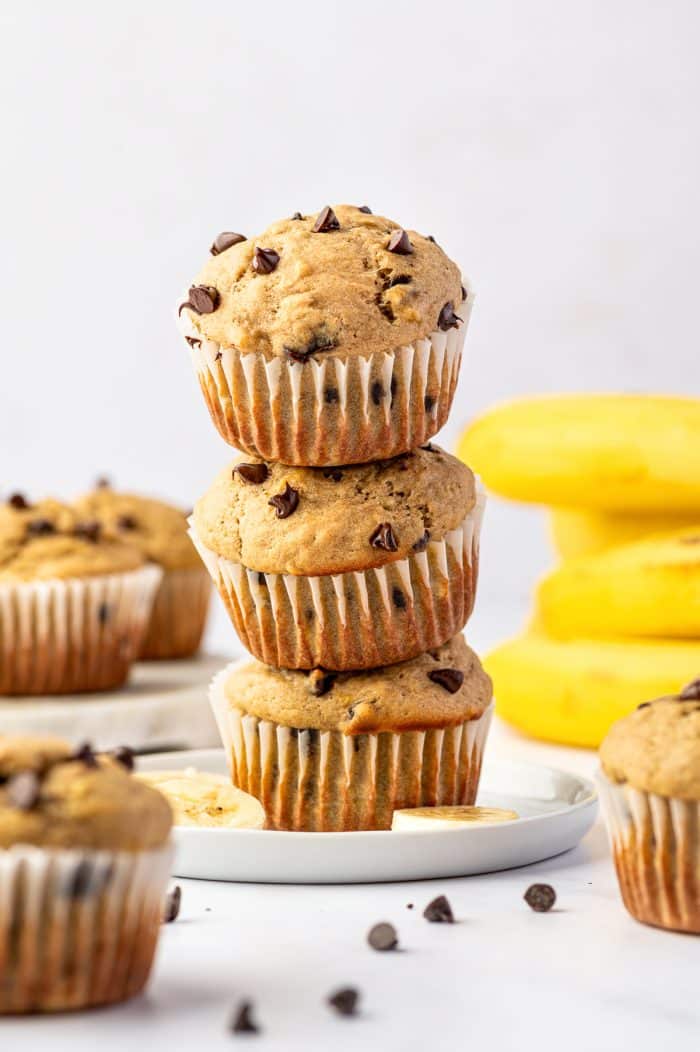 Three golden banana protein muffins with mini chocolate chips stacked on a white plate, surrounded by additional muffins, banana slices, scattered chocolate chips, and whole yellow bananas in the background.