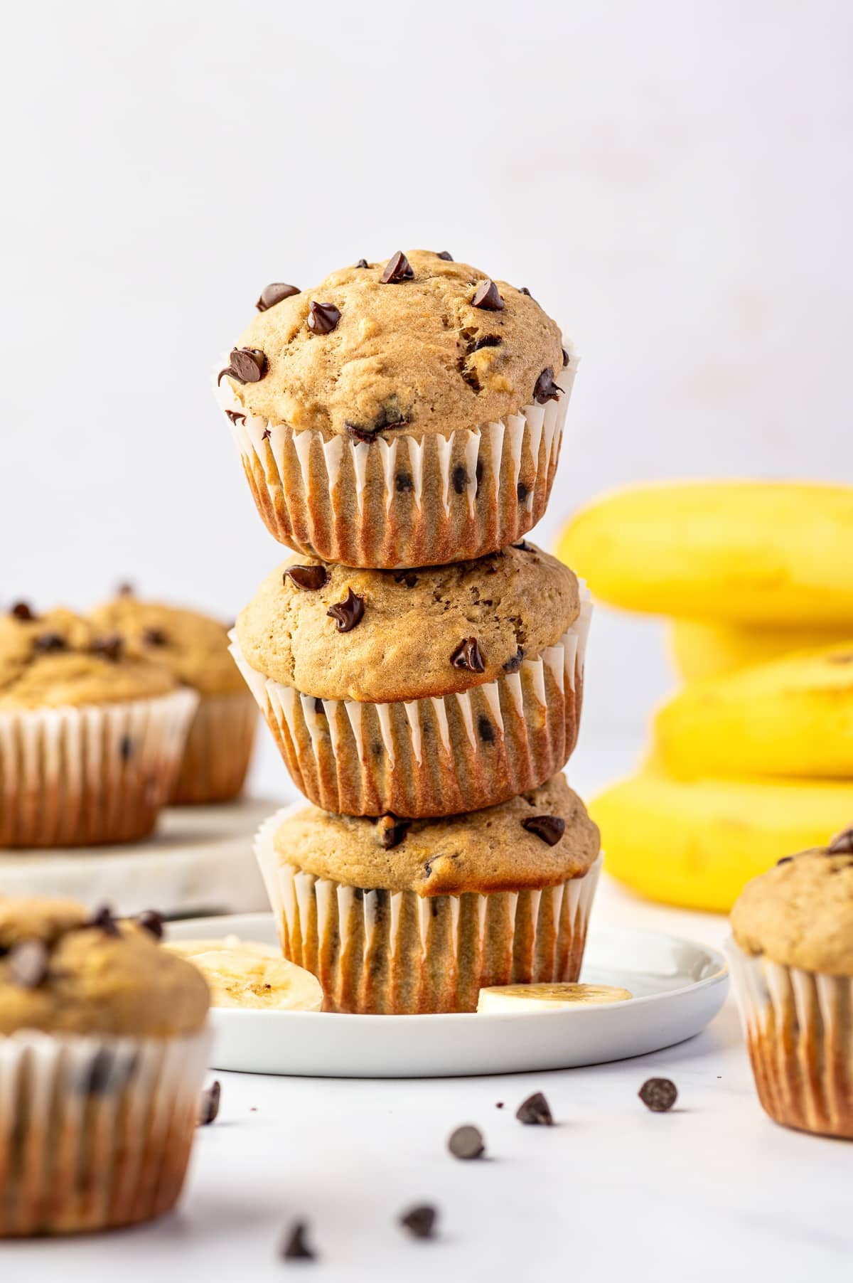 Three golden banana protein muffins with mini chocolate chips stacked on a white plate, surrounded by additional muffins, banana slices, scattered chocolate chips, and whole yellow bananas in the background.