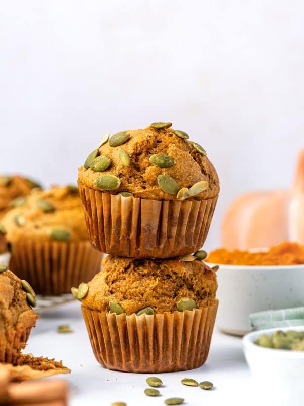 Two golden-brown protein pumpkin muffins stacked on top of one another in brown paper liners and topped with green pepitas. A white bowl of bright orange pumpkin puree, a small white bowl of pepitas, a pale orange pumpkin, and a green striped kitchen towel are softly blurred in the background.