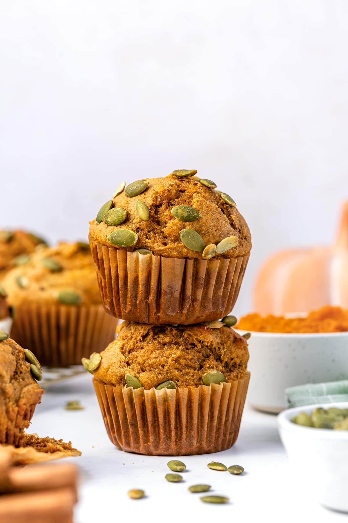 Two golden-brown protein pumpkin muffins stacked on top of one another in brown paper liners and topped with green pepitas. A white bowl of bright orange pumpkin puree, a small white bowl of pepitas, a pale orange pumpkin, and a green striped kitchen towel are softly blurred in the background.