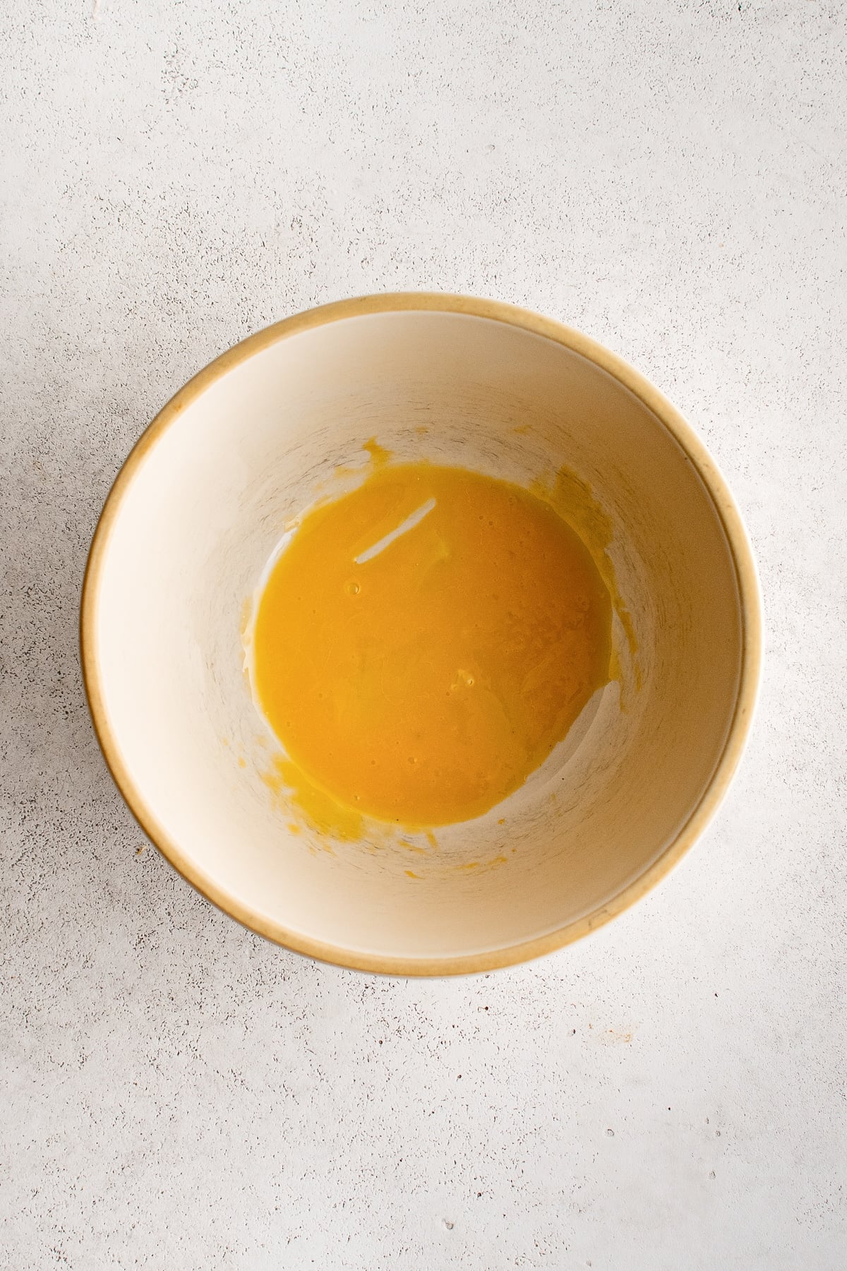 Overhead view of a whisked golden-yellow egg yolk in the bottom of a cream-colored ceramic mixing bowl with a tan rim, set on a light gray textured surface.
