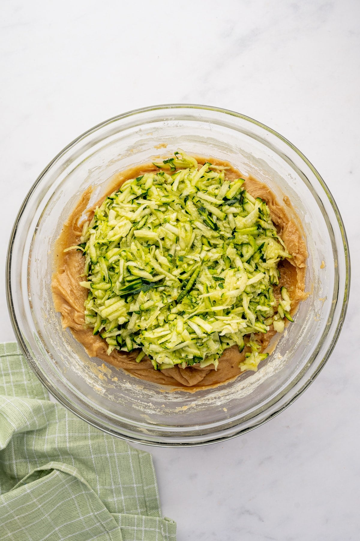 Shredded green zucchini piled over tan zucchini muffin batter in a large clear glass mixing bowl on a white marble surface, with a pale green plaid kitchen towel in the lower left corner.