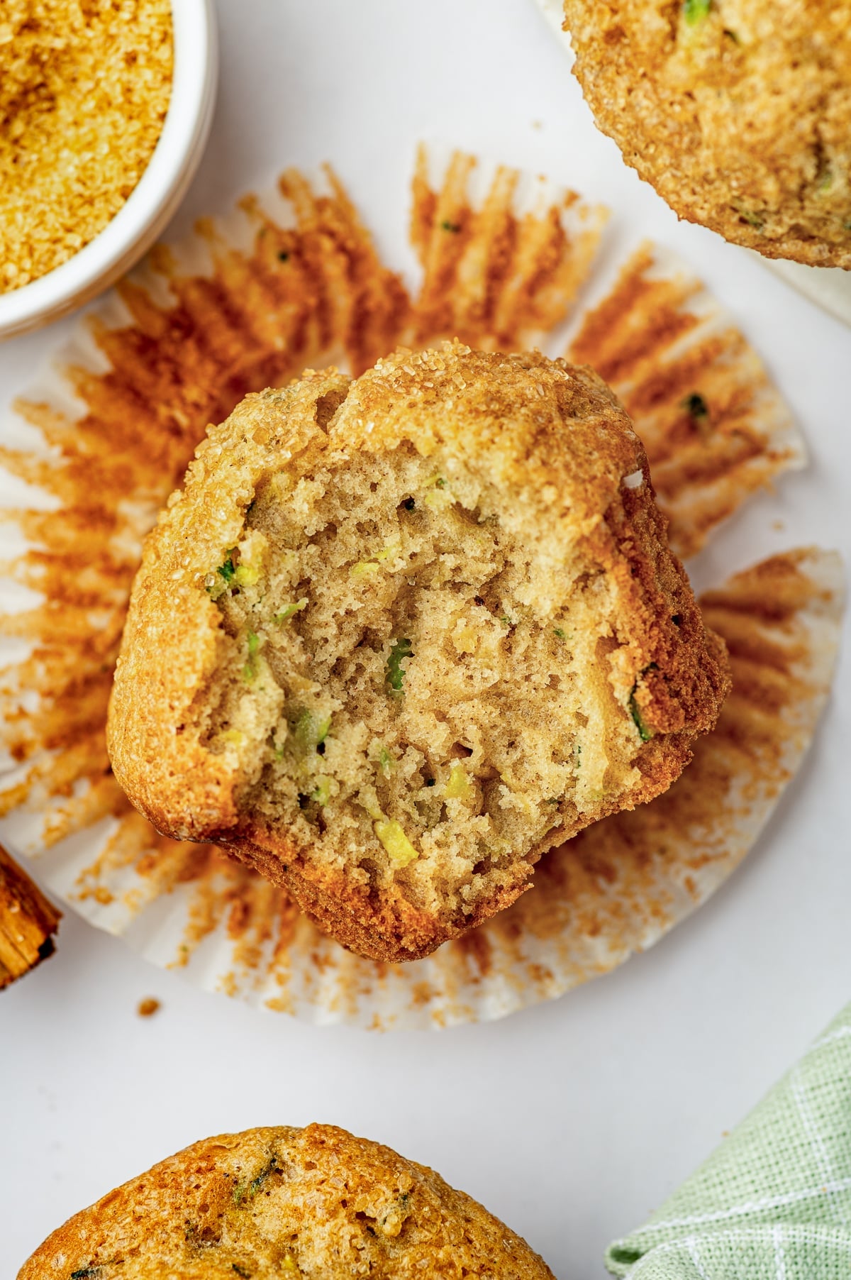 A zucchini muffin broken open to reveal a soft, fluffy golden crumb with small green zucchini pieces, sitting on its unfolded white paper liner. Coarse sugar, another muffin, a cinnamon stick, and a pale green plaid towel surround it on a white surface.