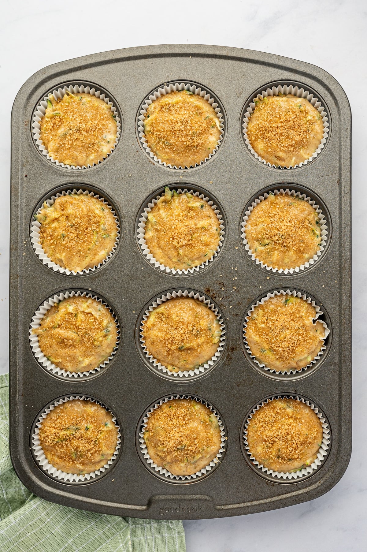 Twelve white paper muffin liners filled nearly to the top with zucchini muffin batter in a dark gray metal muffin pan, each sprinkled with coarse golden sparkling sugar before baking.