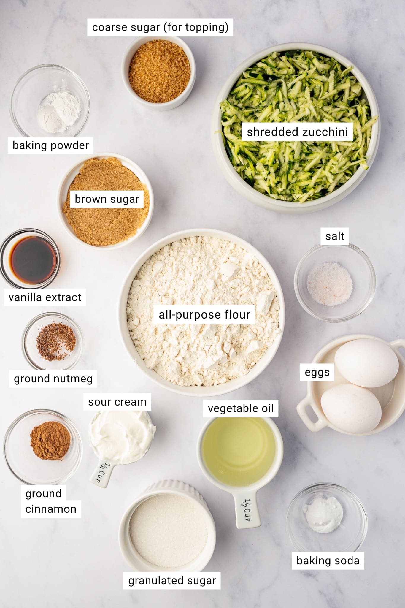 Overhead labeled ingredient layout for zucchini muffins on a light gray marble surface. White bowls and clear glass dishes hold shredded green zucchini, all-purpose flour, brown sugar, granulated sugar, two white eggs, vegetable oil, sour cream, vanilla extract, ground cinnamon, ground nutmeg, baking powder, baking soda, salt, and coarse sugar for topping. Black text on white labels identifies each ingredient throughout the image.