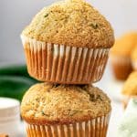 Vertical Pinterest image featuring two golden zucchini muffins stacked on a white surface with tall domed tops and sparkling sugar, styled with cinnamon sticks, dark green zucchini, and a pale green plaid napkin. A white text box at the top reads "Zucchini Muffins" with an orange line underneath, and THEFORKEDSPOON.COM appears along the bottom.