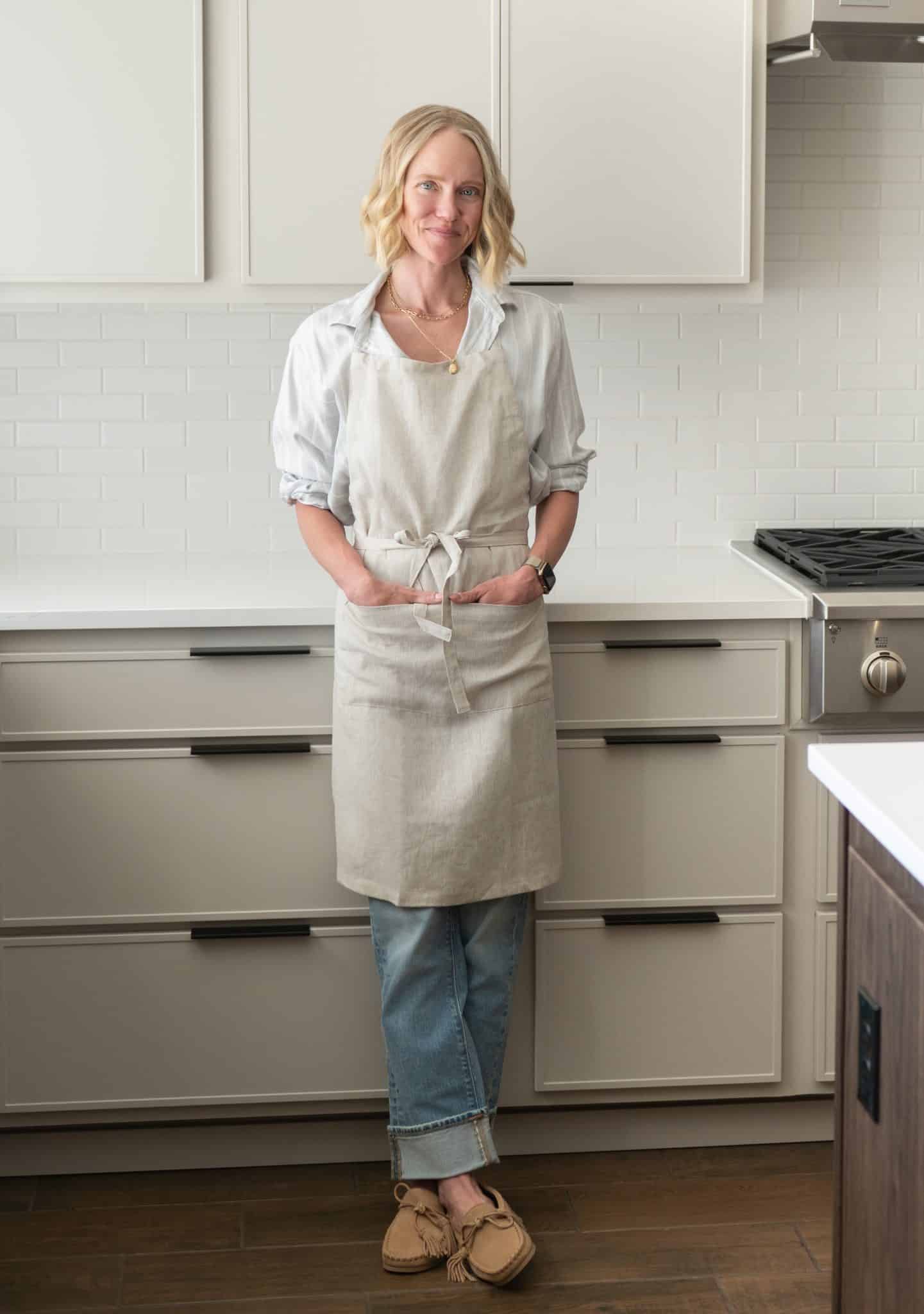 Chef Jessica Randhawa standing wearing a linen apron and standing in her bright home kitchen near stove