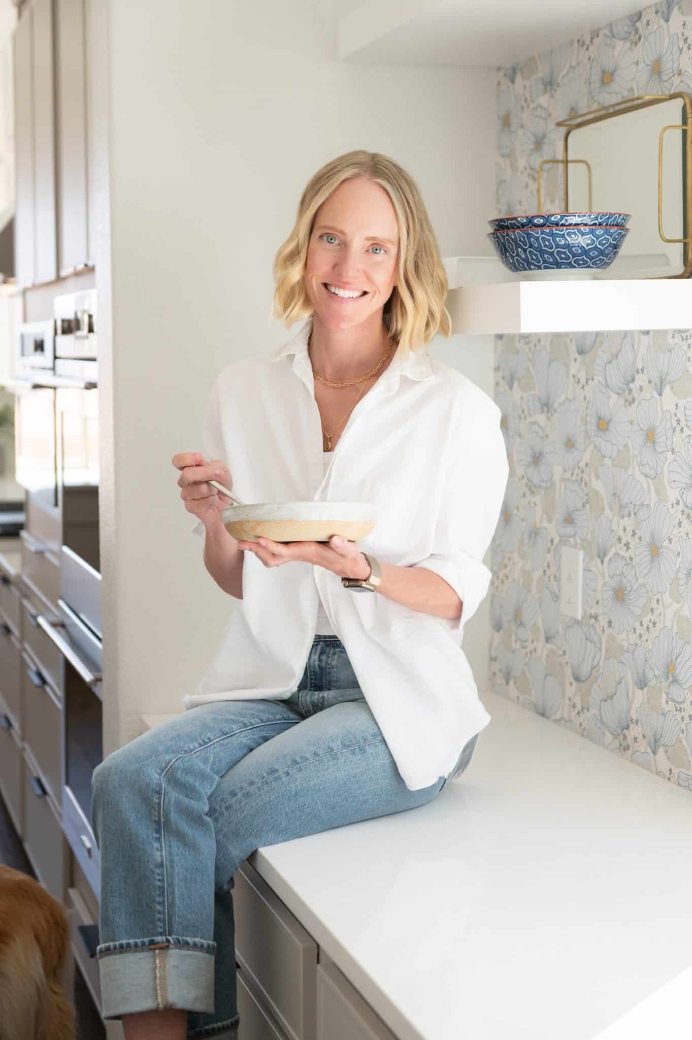 Chef Jessica Randhawa sitting on a kitchen counter holding a bowl of food in her bright home kitchen.