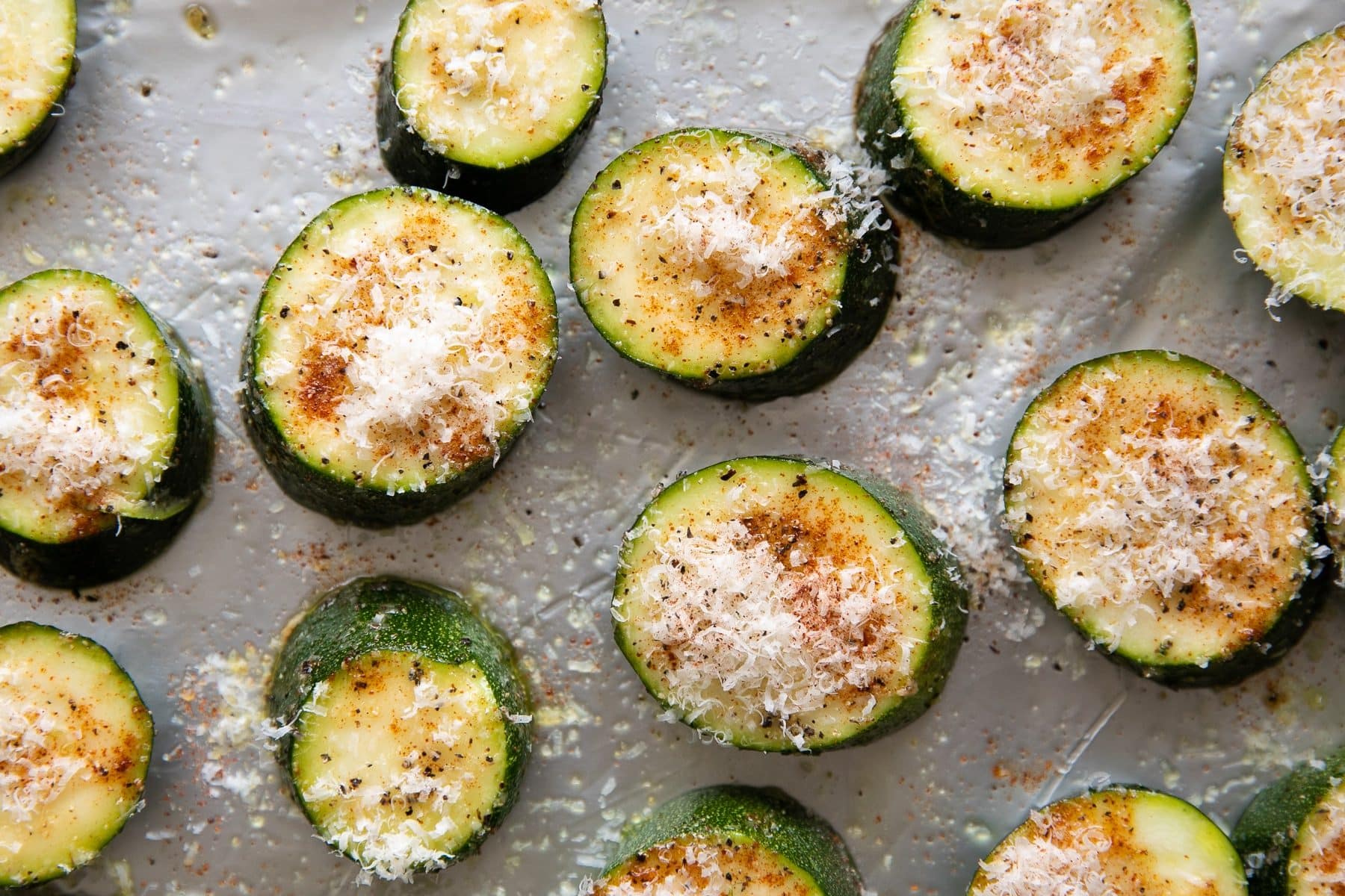 Zucchini slices on a baking sheet seasoned with salt, pepper, paprika, garlic, and topped with grated parmesan cheese.