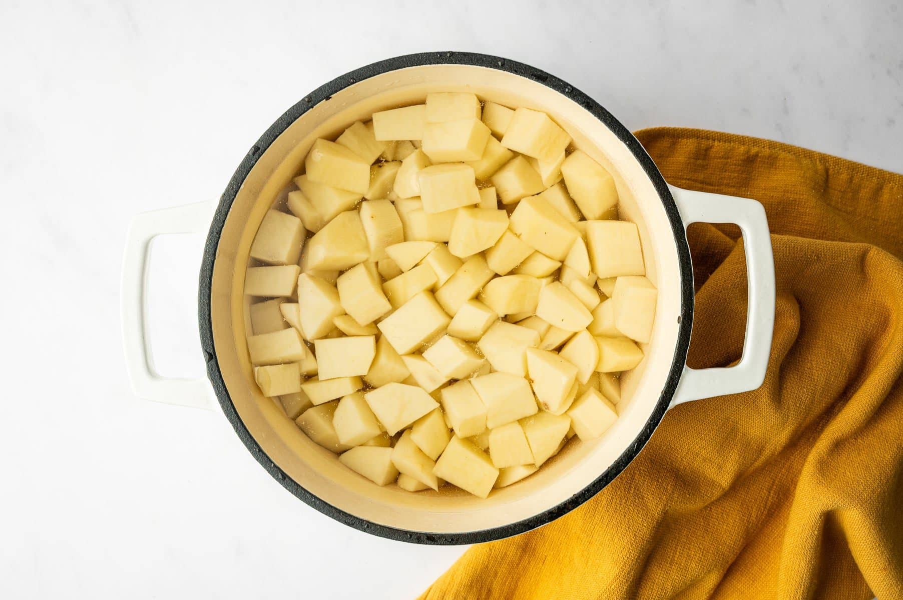 Peeled Russet potatoes cut into 1-inch chunks and covered with cold water in a white enameled Dutch oven with a black rim, photographed from overhead on a white surface with a mustard-yellow kitchen towel underneath.