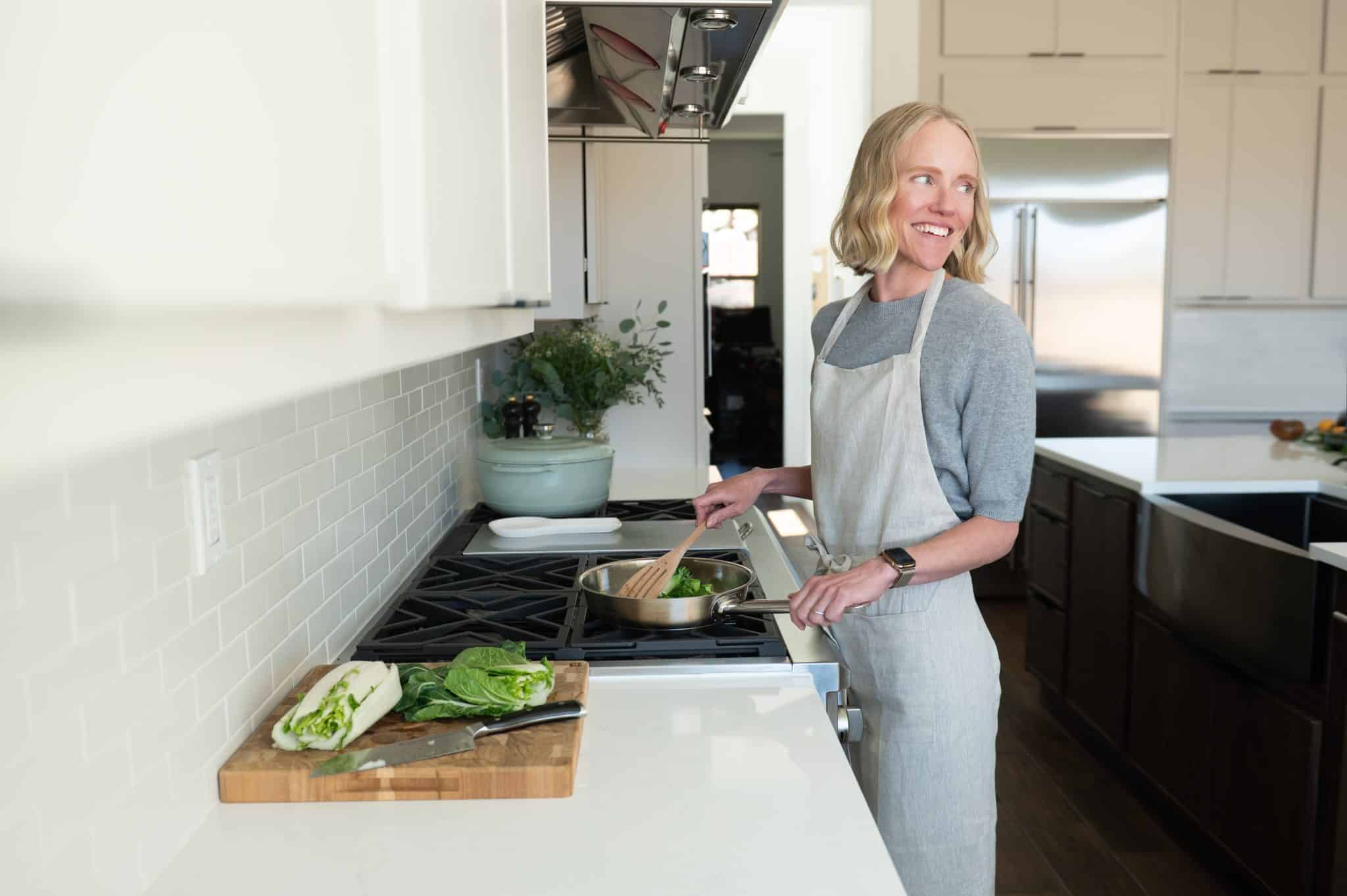 Jessica Randhawa cooking bok choy in a stainless steel skillet on a gas stovetop, wearing a light linen apron in a bright modern kitchen, with fresh bok choy and a chef's knife on a wooden cutting board beside her.