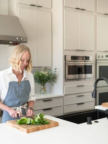 Chef Jessica Randhawa chopping fresh bok choy on a wooden cutting board in her bright home kitchen, wearing a blue striped apron over a white shirt.