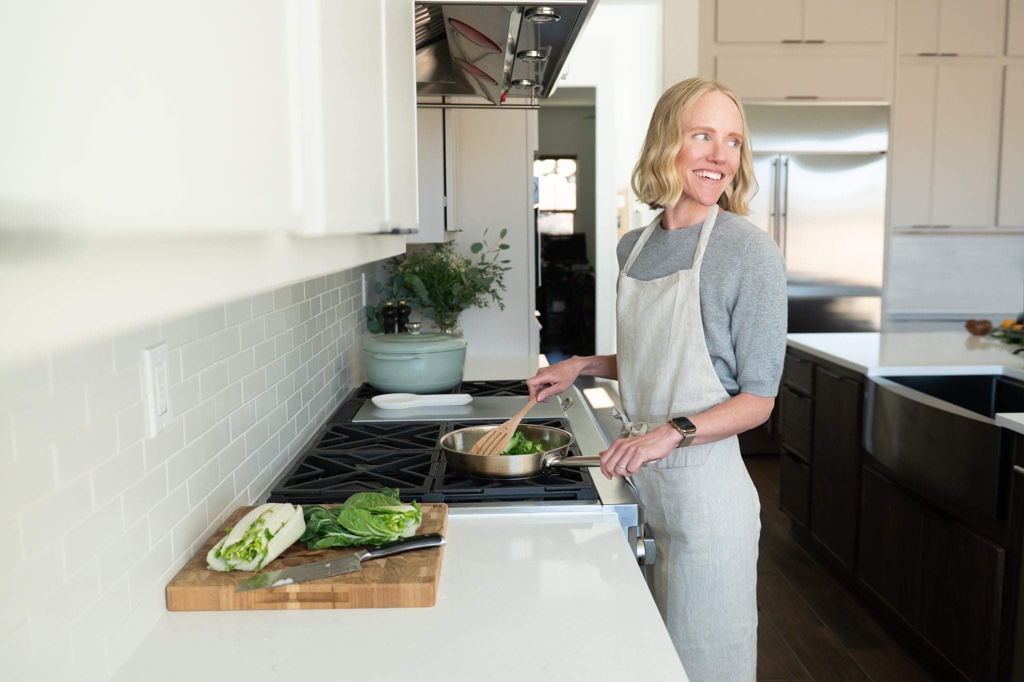 Jessica Randhawa cooking bok choy in a stainless steel skillet on a gas stovetop, wearing a light linen apron in a bright modern kitchen, with fresh bok choy and a chef's knife on a wooden cutting board beside her.