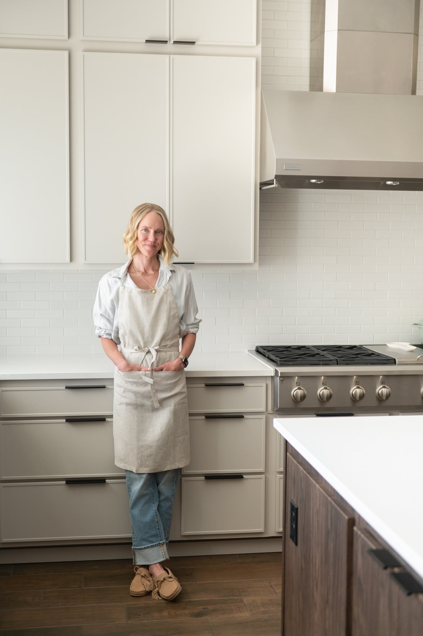 Chef Jessica Randhawa standing wearing a linen apron and standing in her bright home kitchen near stove