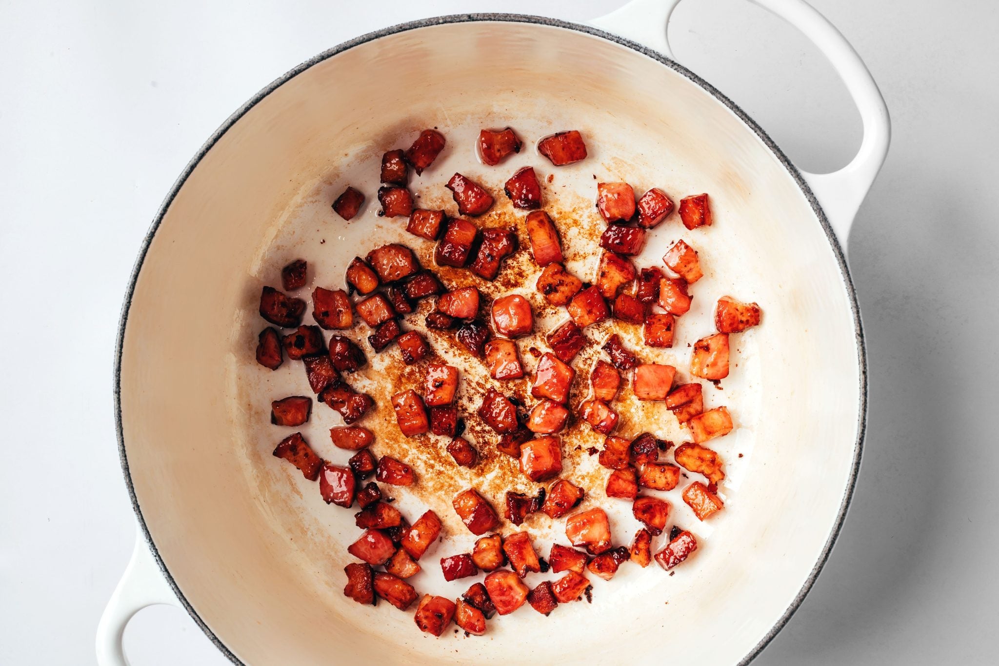 Pieces of thick-cut bacon cooked until browned and crisp in a white Dutch oven, with rendered bacon fat and browned bits remaining in the bottom of the pot.