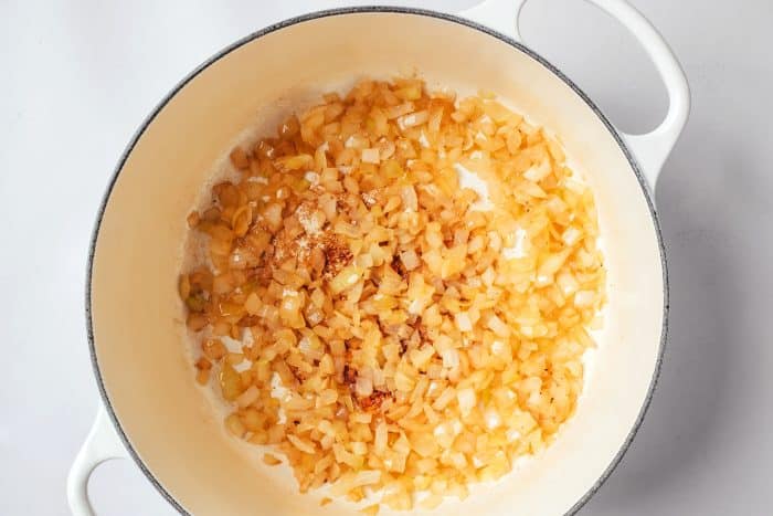 Diced yellow onions softened and lightly golden in a white Dutch oven with browned bits from the bacon and ground beef visible on the bottom of the pot.