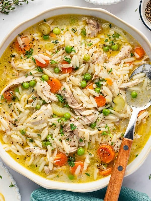 A large, shallow white bowl filled with lemon chicken orzo soup, featuring shredded chicken thighs, tender orzo pasta, sliced carrots, celery, and green peas in a golden broth. The soup is garnished with chopped parsley, fresh thyme, and cracked black pepper. A silver spoon with a wooden handle rests in the bowl, with fresh herbs, lemon wedges, and a teal linen napkin nearby.
