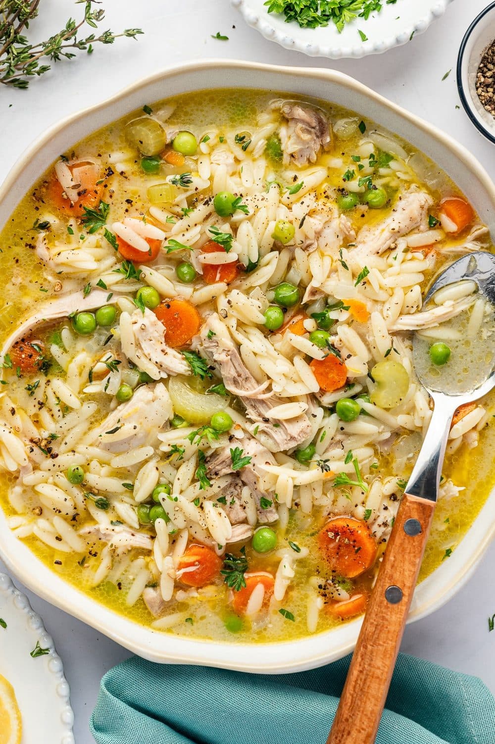 A large, shallow white bowl filled with lemon chicken orzo soup, featuring shredded chicken thighs, tender orzo pasta, sliced carrots, celery, and green peas in a golden broth. The soup is garnished with chopped parsley, fresh thyme, and cracked black pepper. A silver spoon with a wooden handle rests in the bowl, with fresh herbs, lemon wedges, and a teal linen napkin nearby.