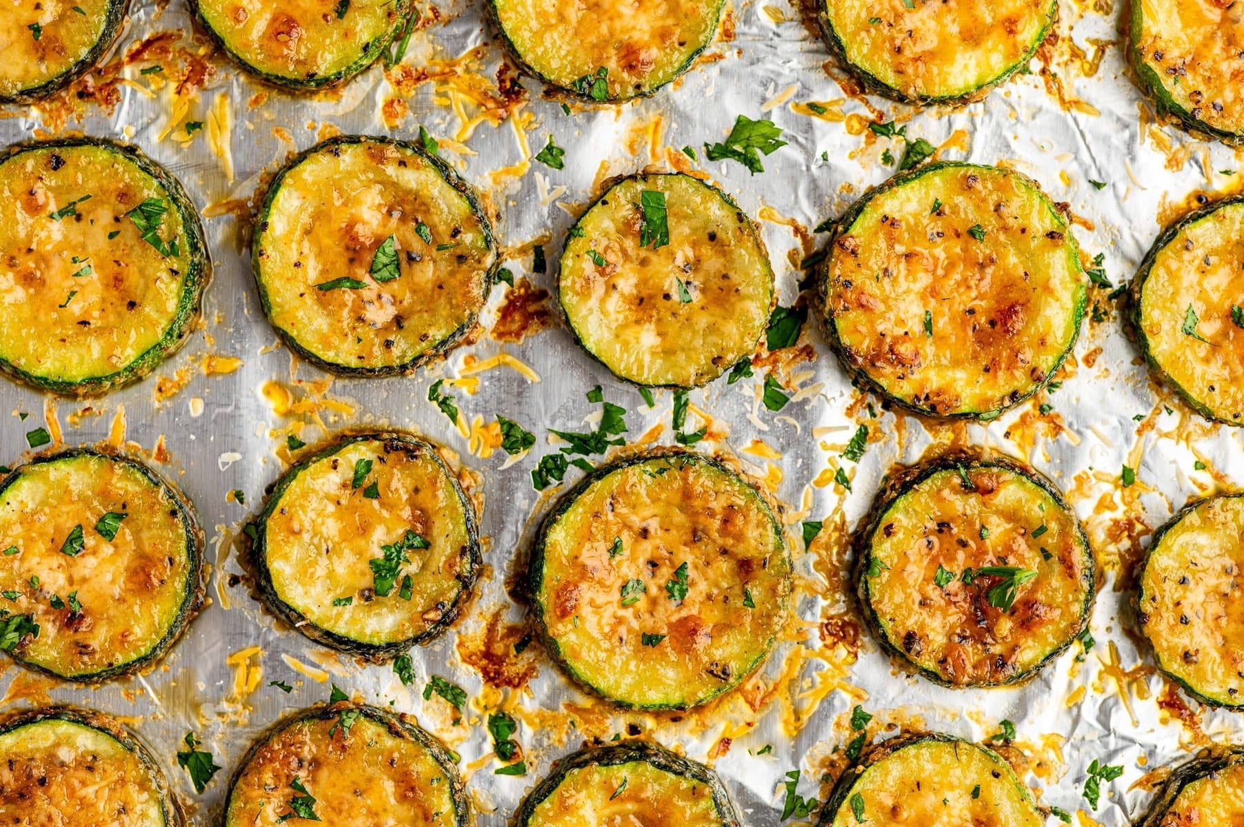 Several thick roasted zucchini rounds resting on crinkled aluminum foil, their dark green skins surrounding tender, golden centers covered with melted Parmesan cheese. Browned cheese spots, coarse black pepper, and chopped fresh parsley are scattered over the zucchini and baking sheet.