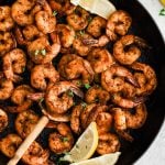 A rustic wooden spoon scoops up perfectly seared shrimp from a pan of blackened shrimp, with steam rising and lemon wedges on the side.