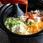 Ingredients for Buffalo Chicken dip being put into the slow cooker, with the hot sauce being poured